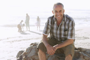 Photo of a man wearing a checkered shirt and shorts. He is sitting on a rock at the beach. The sea is in the background. He is smiling at the camera.